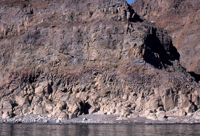 Fig. 51 Lava flow with a basal colonnade of coarse columns along the shore, changing upwards to slender columns in an entablature zone with a wavy column pattern. The overlying part of the flow is thoroughly brecciated. Despite these signs of emplacement into water, the flow is capped by a red-oxidised sediment layer (white arrow) indicating that the top of the flow was above water level after emplacement. Height of field of view c. 15 m. East side of Kigataq island. Photo: Asger Ken Pedersen.
