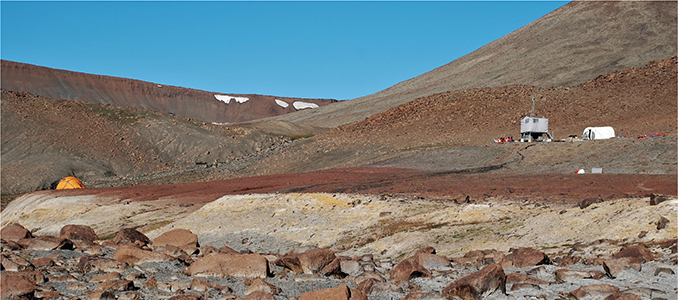 Fig. 5 The Brorson Halvø-1 drill site viewed approximately towards the ENE. The drill-rig stands on the dark grey mudstones of the Stratumbjerg Formation with the well-defined contact to the bright red Rødryggen Member clearly visible below. Downhill, the contact between the Rødryggen Member and the underlying Albrechts Bugt Member (yellow) stands out. The contact to the Storsletten Member (grey), in turn, underlying the Albrechts Bugt Member appears somewhat gradational, but in a clean section, it is very well defined (see Fig. 6; Photo: Annette Ryge).